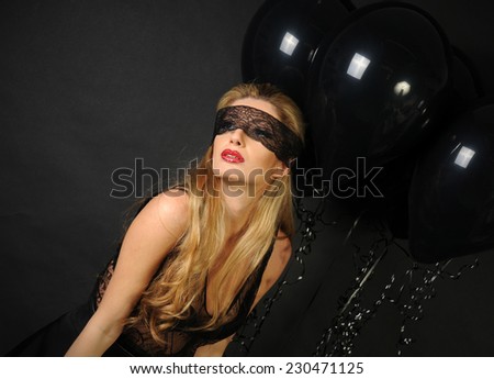 Young beautiful slim girl wearing black lace sensual top and black skirt with evening make up and black lace band on her eyes posing and playing with black balloons isolated on black background