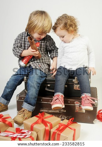two little kids seating on the vintage leather trolleys among beige present packs with red ribbon eating chocolate and looking down intaracting