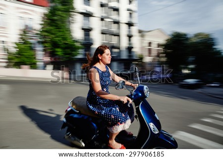Young woman wearing dress is riding the scooter along the street. The photo is made using panning technique, so the natural motion blur is introduced to the image.