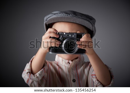 Young boy with a grey corduroy cap and button up striped shirt posing with a camera like a press photographer of old. Shallow depth of field.