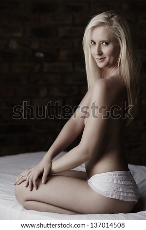 Beautiful and very sexy young adult caucasian woman topless but in white lingerie bottoms with blonde hair and blue eyes, in a bedroom setting with typical boudoir poses