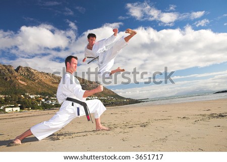 Young adult men with black belt practicing on the beach on a sunny day. The man doing the flying kick in the background has movement. Focus on the standing man
