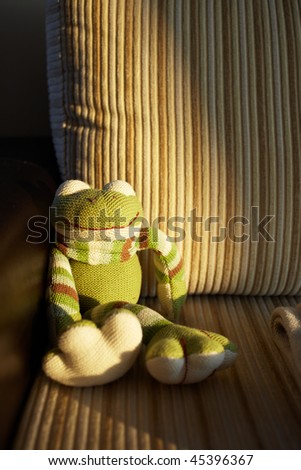 Green handmade knitted toy frog sitting in the last sunbeam of sunset on a brown corduroy and leather couch. Very shallow Depth of Field, natural light.