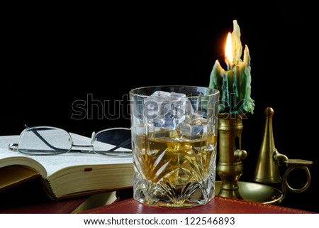A glass of whiskey with ice standing next to an antique book, with candle and glasses