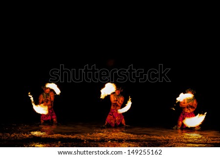 Bathed in an orange glow, three male fire dancers on a Hawaiian beach, stand in the ocean and spin sticks with fire on either side. The glow of the fire is also reflected on the water.