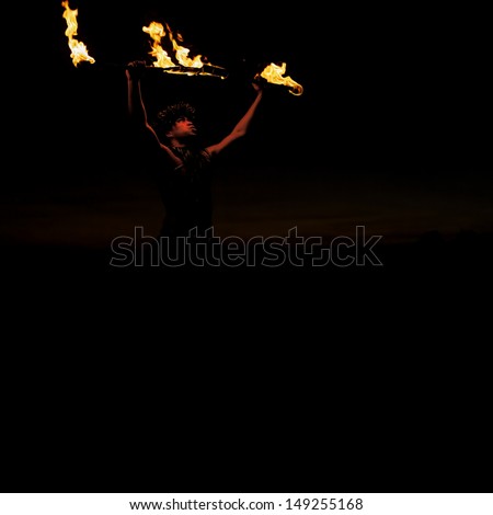 A male, Hawaiian fire dancer is illuminated by the sticks of fire he holds above his head. Square orientation