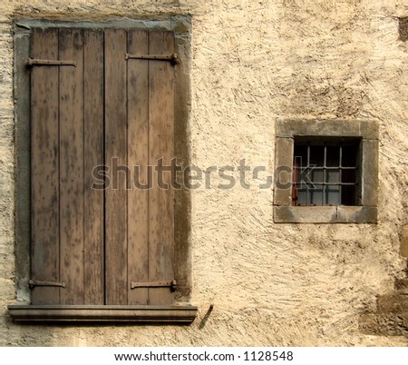 Small window and big closed window with brown shutters. Rough, yellow or beige wall of an old house in a small Italian town or village.