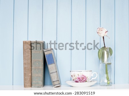Old antiquarian ancient books, china cup and rose in the bottle on bookshelf on the blue wooden background