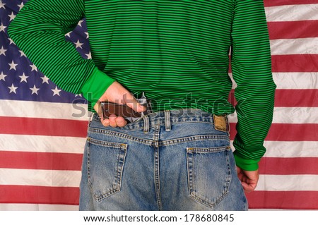 Man standing in front of a US flag holding the grip of a handgun which is stuck behind his back in his pants