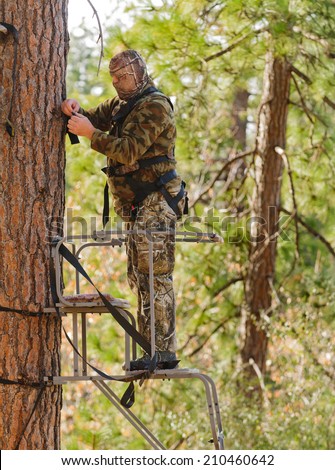 Bow hunter in a ladder style tree stand correctly attaching a fall arrest harness to a strap around the tree
