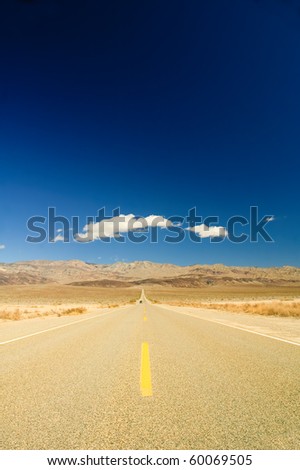 long straight road across Death Valley with a distant motor vehicle coming towards the camera, a deep blue sky late in the afternoon, and a small puffy clouds in the sky