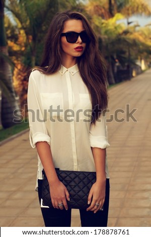 Young fashionable trendy girl posing at tropical alley between palms in evening soft light. Vogue style toned portrait of young woman in black jeans, white blouse and sunglasses holding handbag