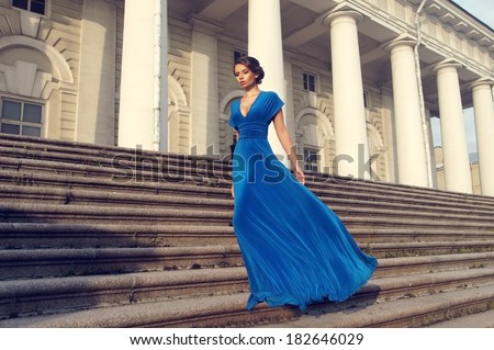 Fashion style portrait. Young elegant woman in blue long flying dress posing at stairway against old city building