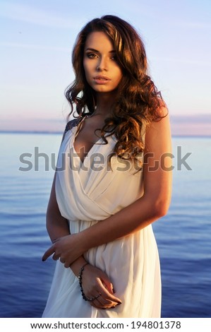 fashion portrait of young beautiful sexy lady in white dress with decollete posing against blue sea and sky at sunset