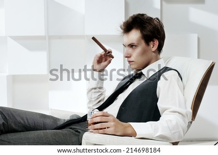 Fashion portrait in modern white office interior. Young handsome man sitting in armchair with cigar and glass of whiskey