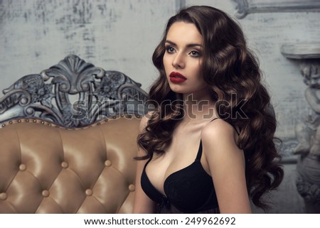 Fashion portrait of young beautiful sexy woman with long wavy hair. Pretty girl sitting in black bra or lingerie in luxury interior.