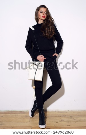 Young stylish girl posing in black costume trousers and pullover and black boots with white handbag in simple interior against white wall