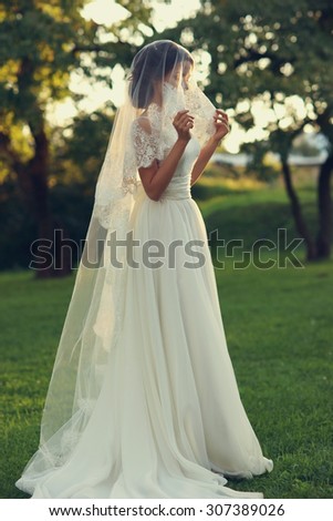 Wedding. Young beautiful bride in white dress and veil standing at green lawn between trees at sunset. Summer full length atmospheric portrait portrait. Side view