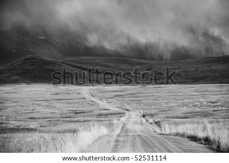 Black-and-white image of storm cloud over barren landscape with mountains in the background. A dirt road recedes across Montana's National Bison Refuge. Horizontal shot.