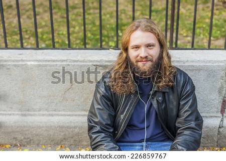 Blond long hair and beard young adult hipster man listening music. Outdoor, urban scene.