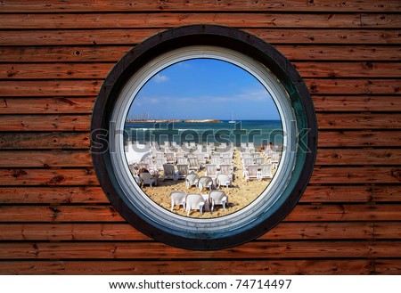 Round window of the wooden house with view on a lot not occupied beach chairs on perfect beach of the Mediterranean sea