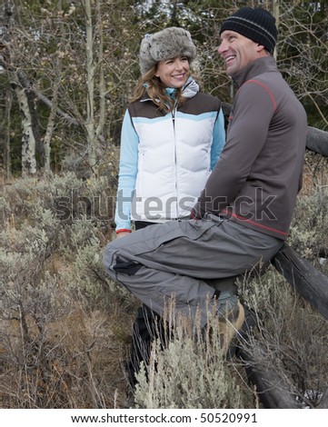 A couple stands together by a rustic wooden fence in a wooded area. They are wearing winter clothing and smiling. Vertical format.