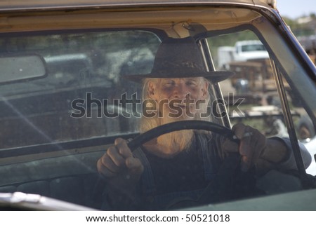 Portrait of an elderly man with a white beard and cowboy hat driving a pickup truck and looking out the windshield at the camera. Horizontal format.