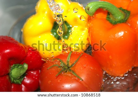 A variety of vegetables, including peppers and a tomato, in a stainless steel sink being washed under a stream of water.