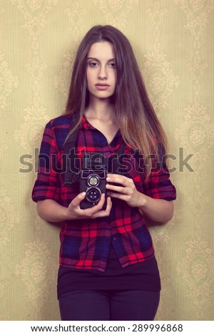Old vintage film camera in the hands of a young girl in a red plaid shirt standing near the old yellow wall.