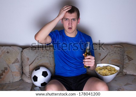 shocked supporter in blue uniform sitting on the sofa and watching football with beer and chips