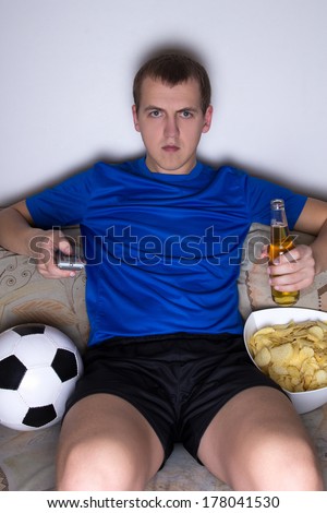 bored man in football uniform sitting in living room and watching tv with beer and chips