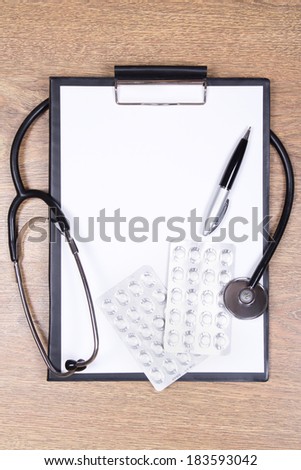 stethoscope, blank clipboard, pen and pills on wooden background