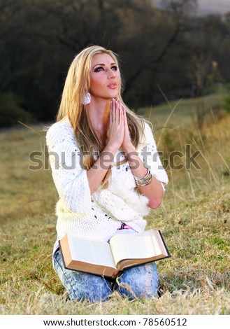 A young blonde lady is sitting on her knees, kneeling and praying with the Bible open on her lap
