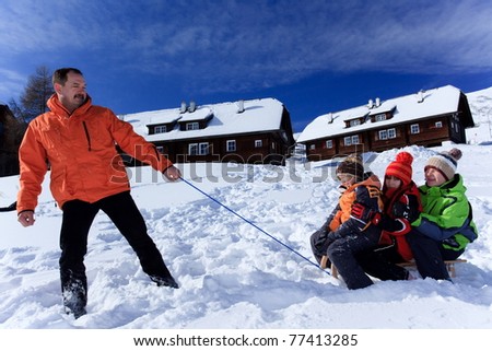 Middle aged father pulling three children on sledge on snowy hillside with houses in background.