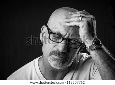 Black and white head shot of a middle aged bald man with facial hair and glasses rubbing his head with his hand on a black background