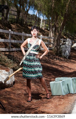 Full length color shot of a young woman in a vintage dress standing on a country roadside with a black and white electric guitar and vintage luggage