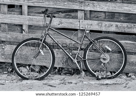 An abandoned bike leaning against a wooden fence. The photo is in black and white.