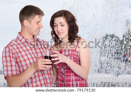 Young man treats his girlfriend to a cool drink. In the background, splash fountain. Hot summer.
