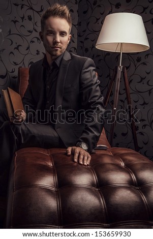 Handsome young man in dark suit with book relaxing on luxury sofa.