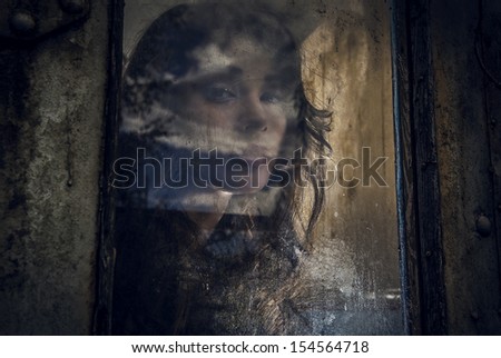 Art portrait of a beautiful young spooky woman, looks through grunge styled rainy window.