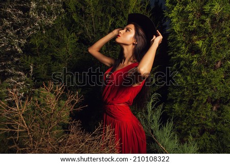 Beauty brunette women in red dress & hat pose at night park.