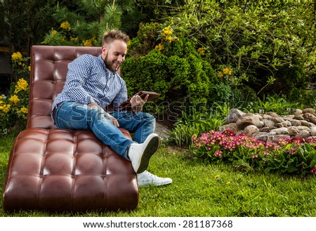 Young handsome man in casual clothes sit in luxury sofa with iPad in summer garden.