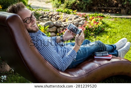 Young handsome man in casual clothes & glasses sit in luxury sofa in summer garden.