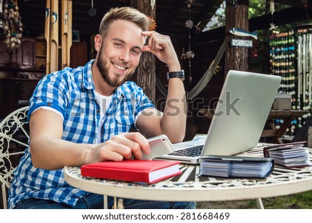 Young smiling handsome man in casual clothes sits at an iron table with computer against country arbor.