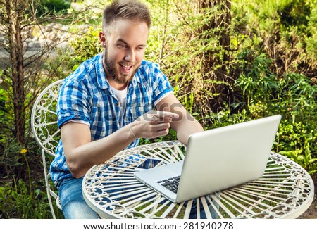 Young smiling handsome man in casual clothes work at an iron table with computer against country garden.