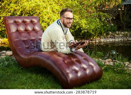 Young handsome man in sit in luxury sofa with iPad in summer garden.
