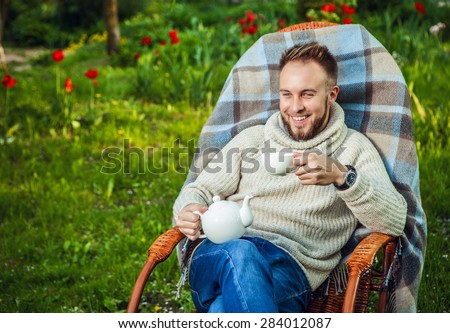 Handsome man relax in rocking-chair with teapot & cup in summer garden.