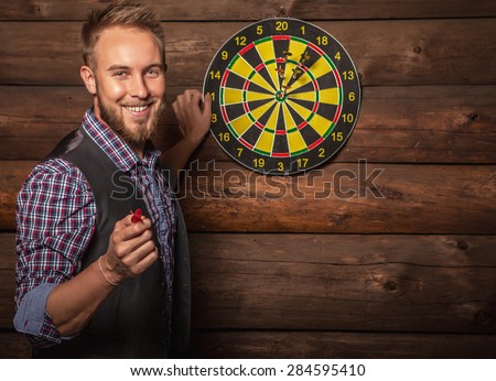 Portrait of young friendly lucky man against old wooden wall with darts game. Concept: Hit in purpose. Photo.