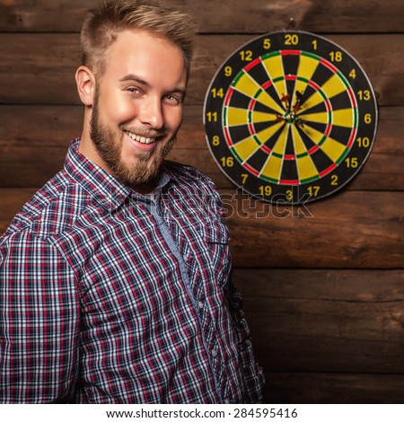 Portrait of young friendly lucky man against old wooden wall with darts game. Concept: Hit in purpose. Photo.