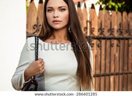 Outdoors portrait of beautiful young girl in luxury white dress posing near gate to country house in sunny summer day.